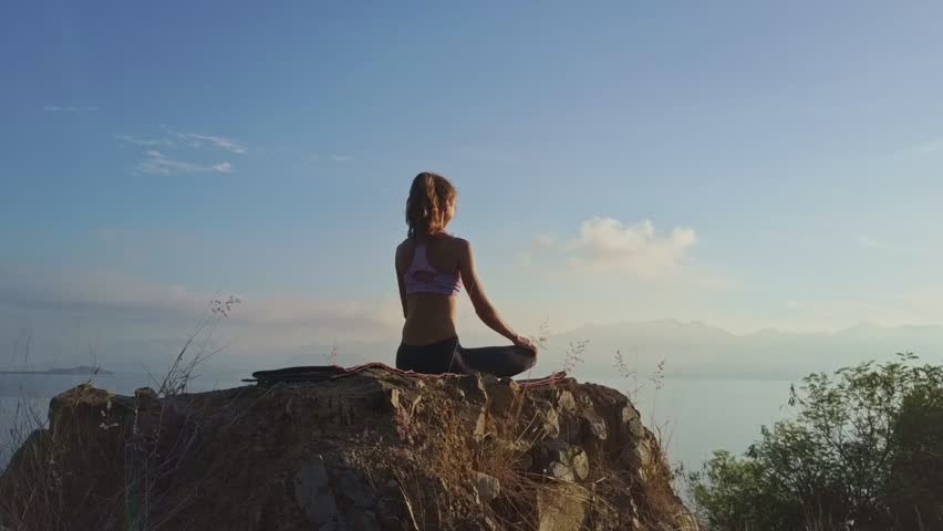 flycam shows backside view young athletic woman sits in Padmasana on rock against large bright morning sun