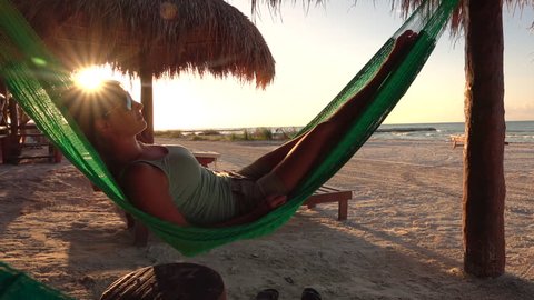 SLOW MOTION Happy young woman on vacation laying and resting in hammock at golden sunset in hot summer on Mexico beach. Serene woman relaxing in swinging hammock on sandy beach at sunrise golden light