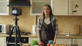 Young attractive woman in apron shooting video food blog about cooking on dslr camera in kitchen