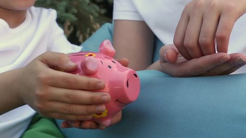 Mother and daughter putting coins into a pink piggy bank sitting on cushions in the park on a summer day.