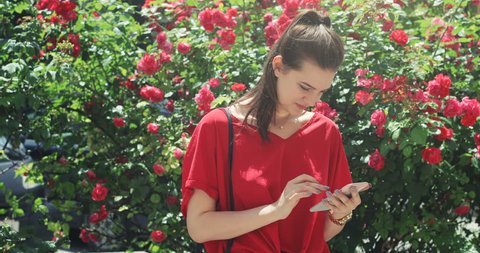Youthful girl in red t-shirt using her smartphone and smiling in street bunch of roses background 4k