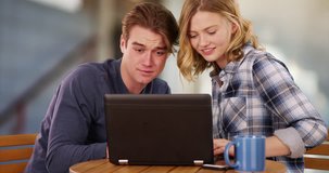 Portrait of two friends sitting indoors and watching something on laptop, laughing and smiling. Happy male and female enjoying each other's company looking at video together on computer. 4k