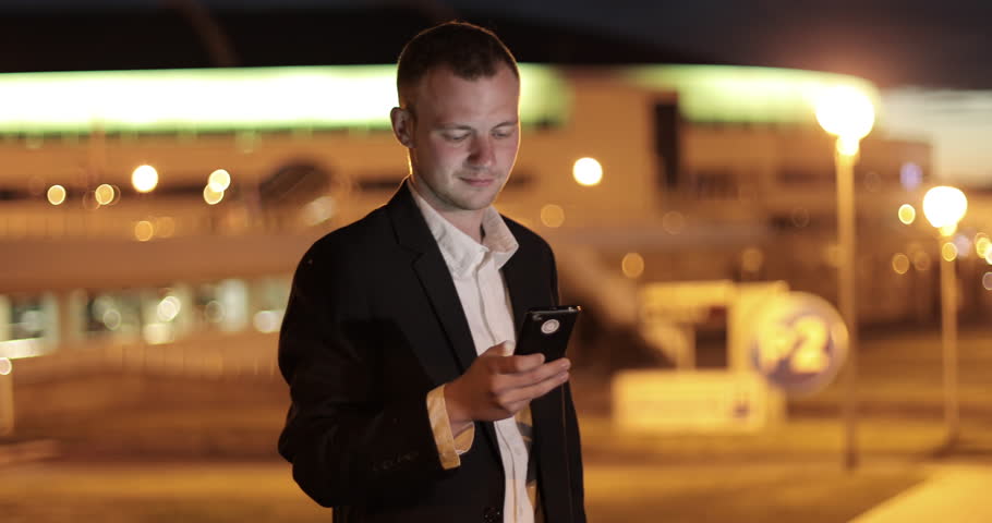 Handsome young business man using mobile phone smiling happy wearing suit jacket outdoors. Man sms texting using app on smart phone at night in city. 4K footage.