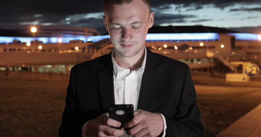 Man sms texting using app on smart phone at night in city. Handsome young business man using smartphone smiling happy wearing suit jacket outdoors. 4K footage.