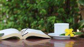 Open book and white coffee cup with eyeglasses on table and fresh nature green garden background