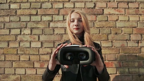 A stylish young girl near a brick wall brings virtual glasses to the camera.