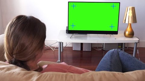 Young Woman Watching TV Relaxed At Home, Panning Shot. Young woman watching television green screen. Panning shot behind models shoulders
