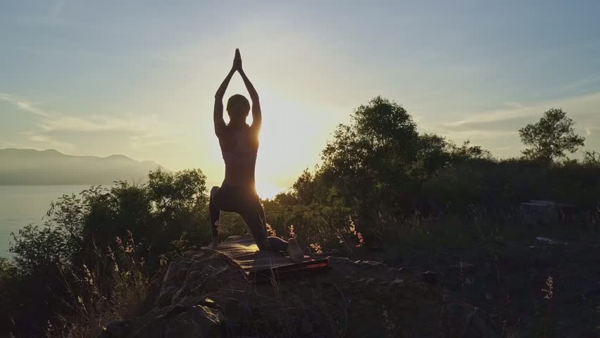 backside view young athletic woman stands in Virabhadrasana on rock against large bright morning sun