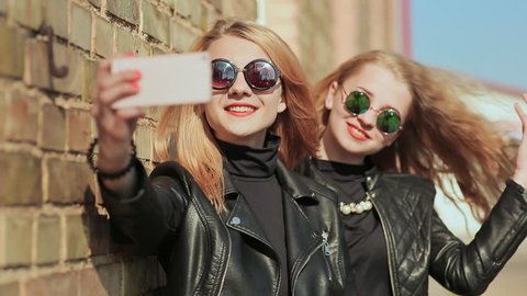 Two girlfriends in sunglasses and stylish leather jackets take a photo from a mobile phone near a brick wall on the street. Do selfie.