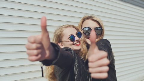 Two smiling stylish girls in leather jackets and sunglasses show a gesture of thumbs up. Background of white horizontal rolling shutters.