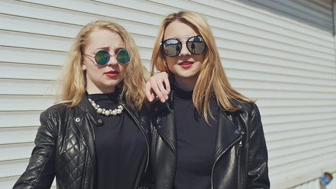 Two stylish young girls in leather jackets and sunglasses on a white background show a gesture. Finger down. Background of white horizontal rolling shutters.
