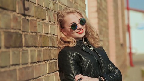 Stylish young blonde in sunglasses and black leather jacket near a brick wall on the street.