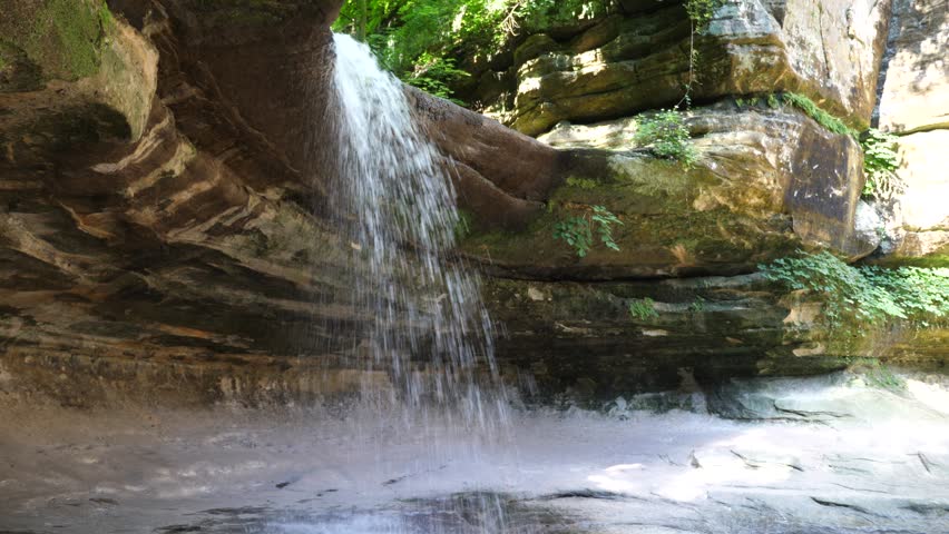 Waterfall flowing over a limestone overhanging cliff in the LaSalle canyon at Starved Rock State Park in Illinois with green foliage above and sound of water flowing down to rocky stream bed below