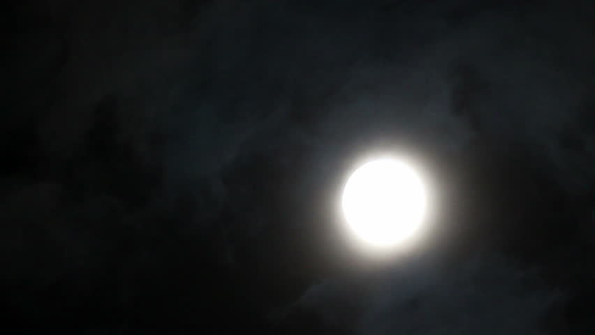 Dark clouds slowly moving through the full moon, shining on a black sky