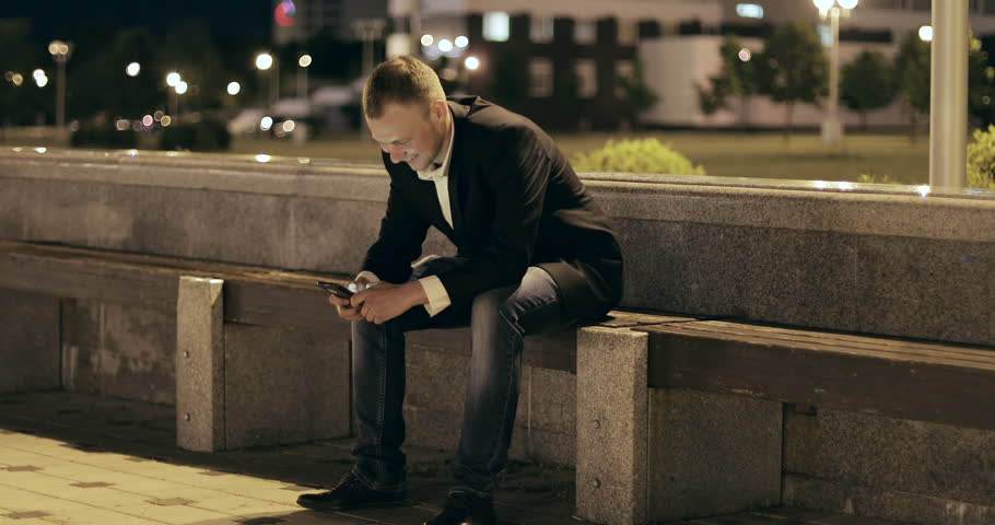 Man sms texting using app on smart phone at night in city. Handsome young business man using smartphone smiling happy wearing suit jacket outdoors. 4K footage.