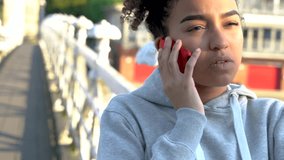 4K video clip of beautiful mixed race African American girl teenager young woman on a bridge over a river, talking on a mobile cell phone 