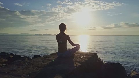 drone moves around girl sitting in pose ArdhaPadmasana against unforgettable sun reflection on ocean surface