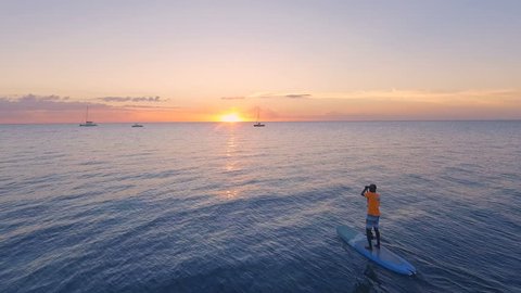 AERIAL: Stand up paddling in the Indian Ocean against the background of an orange sunset near the coast of Africa. African man on SUP board. 4K.