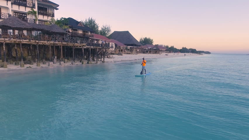 AERIAL: Stand up paddling in the Indian Ocean against the background of an orange sunset near the coast of Africa. African man on SUP board. 4K.