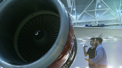 In a Hangar Aircraft Maintenance Engineer Shows Technical Data on Tablet Computer to Airplane Technician, They Diagnose Jet Engine Through Open Hatch. They Stand Near Clean Brand New Plane. 4K UHD.