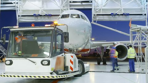 Time-Lapse of a Aircraft Maintenance Hangar Where New Airplane is Toed by a Pushback Tractor/ Tug onto Landing Strip. Crew of Mechanics, Engineers and Drivers Works Busily. Shot on RED EPIC-W 8 Camera
