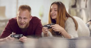 Young cheerful couple lying on sofa teasing each other while playing video games
