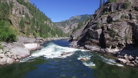Short aerial clip where camera lowers slowly over the rapids of Salmon River in Idaho. A rocky canyon, tall pine trees, and a beautiful blue sky make up the majestic backdrop for this shot.