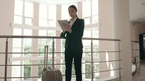 Business woman using her digital tablet and looking on it while sitting on bed near suitcase at the hotel lobby or airport VIP class.