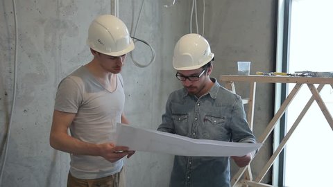 Two men employees considering repair plan standing indoors. They keep in their hands detailed blueprint of finishing works on paper, carefully study it and confer with each other, looking around room