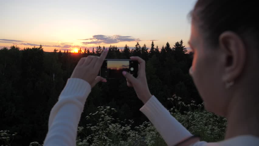 View from the back and side of the woman, shoots and photographs using a smartphone sunset over the forest.  It stands on a hill from where there is a view and the rays of the setting sun are falling.