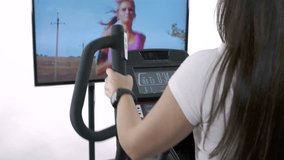 Woman wearing smart watch exercising on elliptical trainer in front of the TV. Television screen displaying video of young girl jogging.
