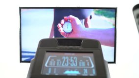 Point of view of elliptical cardio workout in front of the television. TV screen displaying fitness motivation video. Hand with stopwatch on starting line, running fit girl, path through the forest.