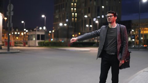 Young handsome businessman in glasses hitchhiking in a night street. He is wearing glasses and a suit. Locked down real time medium shot