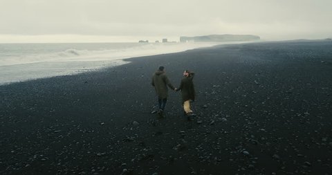 Back aerial view of the young couple walking on the black volcanic beach in Iceland. Romantic date near the sea.