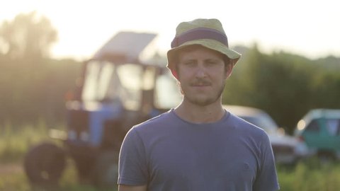 Portrait of young farmer in hat standing at field of organic farm with the tractor on the background. Man looking at camera and smiling in beautiful sunset light.