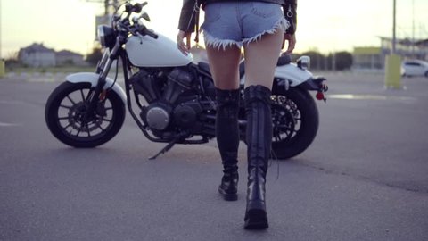 Back view of a female biker in shorts coming up and getting on the motorcycle. Young sexy curly woman in sunglasses on the chopper