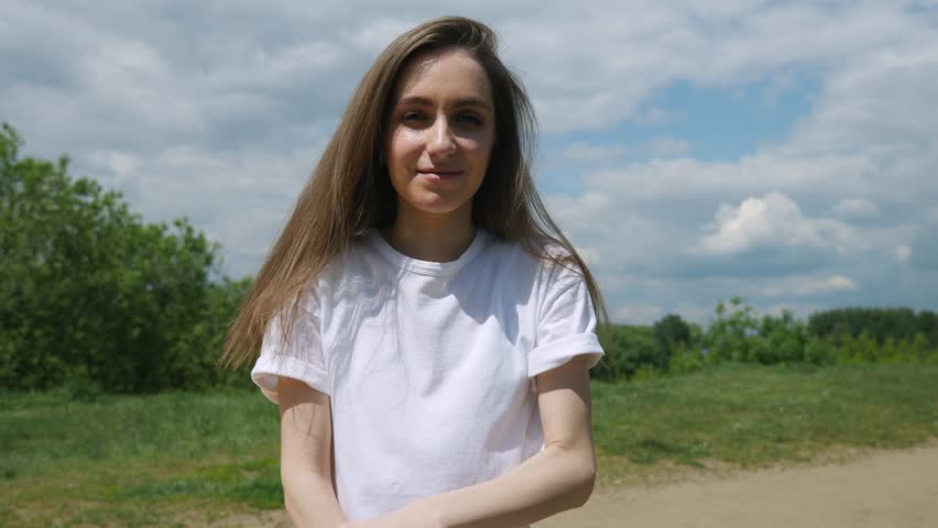 Close up portrait of young girl with long hair in white t-shirt, turning head and looking in camera, smiling. Summer sunny day 4K video.