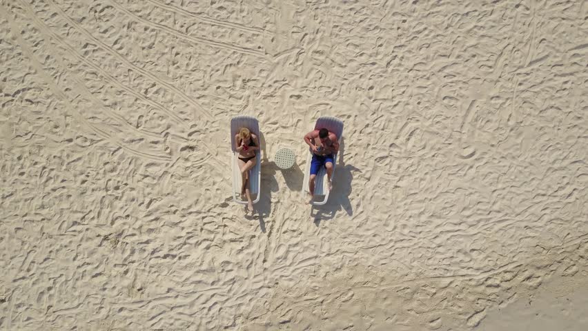Aerial drone camera ascending and rotating high above the couple in love lying in the sun on beach bed deck chairs and drinking cocktails at empty tropical white sand beach on atoll island resort with Royalty-Free Stock Footage #28715236