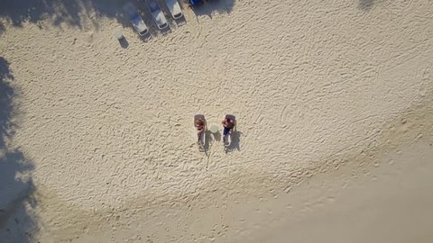 Aerial drone camera ascending above the couple in love lying in the sun on beach bed deck chairs and drinking cocktails at empty tropical white sand beach on atoll island resort with palm trees