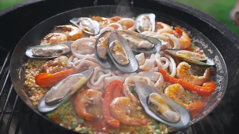 The cook makes paella on the open fire in the big hot pan, grill anf barbecue, spanish food, paella with seafood, food cooking outdoors