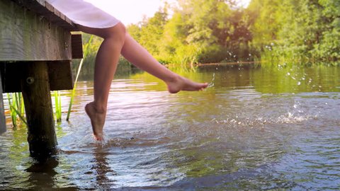 Woman sitting on the edge of a wooden jetty at lake, feet near the water surface, slow motion.
