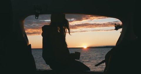 CINEMAGRAPH - seamless loop. CU Silhouette of young Caucasian female sits on a SUV trunk and enjoying hot drink on a lake during sunset. 4K UHD