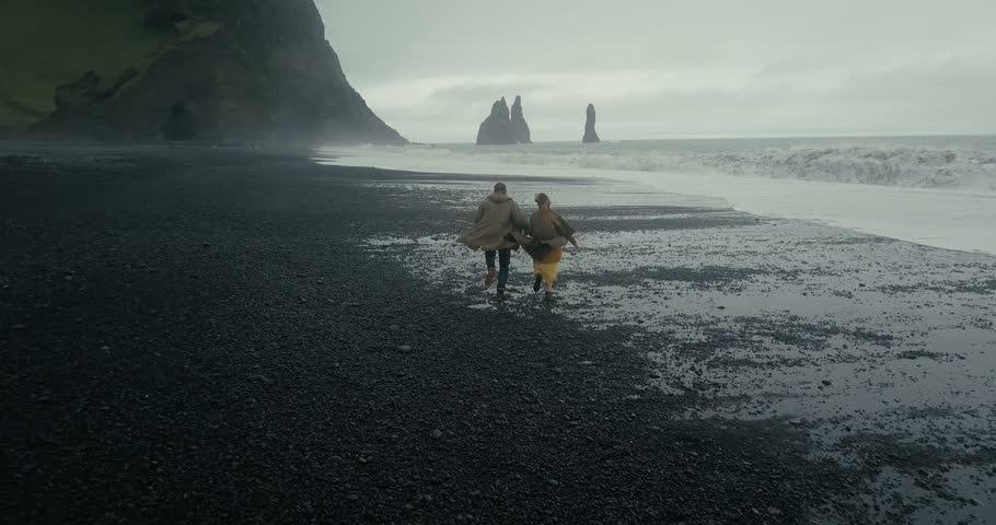 Back aerial view of young hipster couple running on the black volcanic beach near the troll toes in Iceland. Royalty-Free Stock Footage #28728346