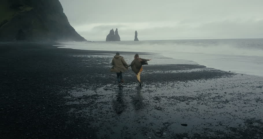 Back aerial view of young hipster couple running on the black volcanic beach near the troll toes in Iceland.