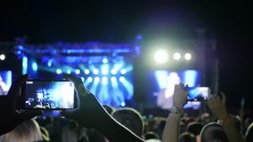 crowd raising hands and shooting video at mobile phone, slow motion, audience at concert or party, lit scene by colourful illumination during night rock festival, many lights on stage