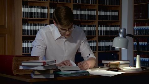 A young serious man with glasses sitting at a table in the library studying and reading books