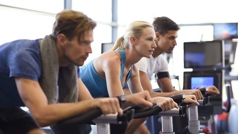 Fitness happy woman on stationary bicycle doing spinning at gym. Fit young woman working out on bike with man. Smiling girl exercising with group of people and looking at camera.
