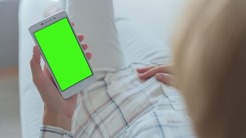 Young Woman in white jeans laying on couch holds SmartPhone with pre-keyed green screen. Perfect for screen compositing