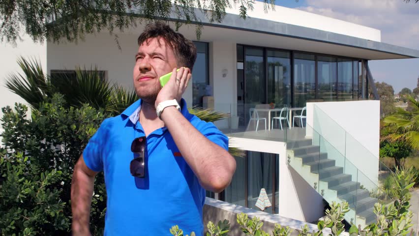 Handsome man in hot weather standing on the terrace of his Villa, and talking on the phone. The sun is shining brightly and he wears glasses