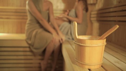 A wooden bucket with spoon on blurred background with girls relaxing in sauna
